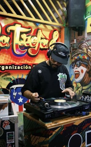Frutamannn DJ tocando en vinilo en La Troja Barranquilla con mural de carnaval de fondo